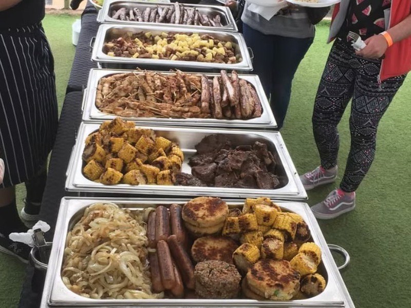 A buffet line of trays with sausages, chicken skewers, steak, corn on the cob, onions and patties