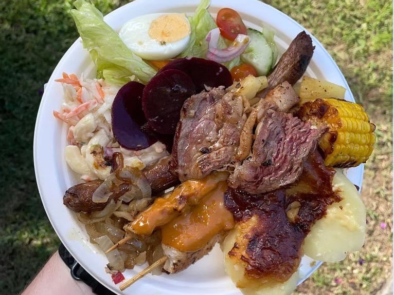 A full plate of steak, sausage, satay chicken skewer, potato bake, corn, beetroot and salads