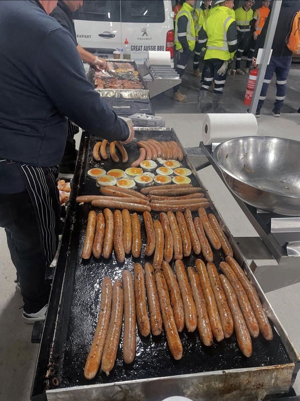Rows of sausages and fried eggs on a big mobile BBQ hotplate at a work site